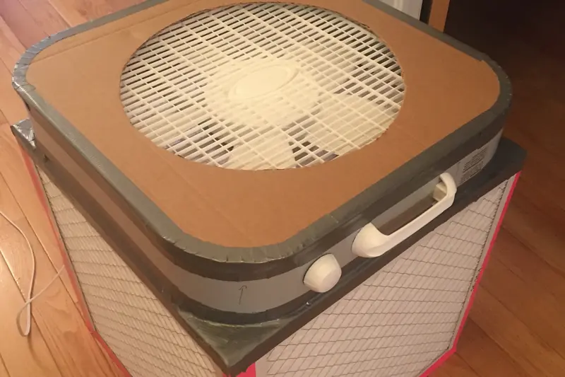 A homemade air cleaner made from a box fan resting on top of a taped cube of pleated furnace filters on a wooden floor
